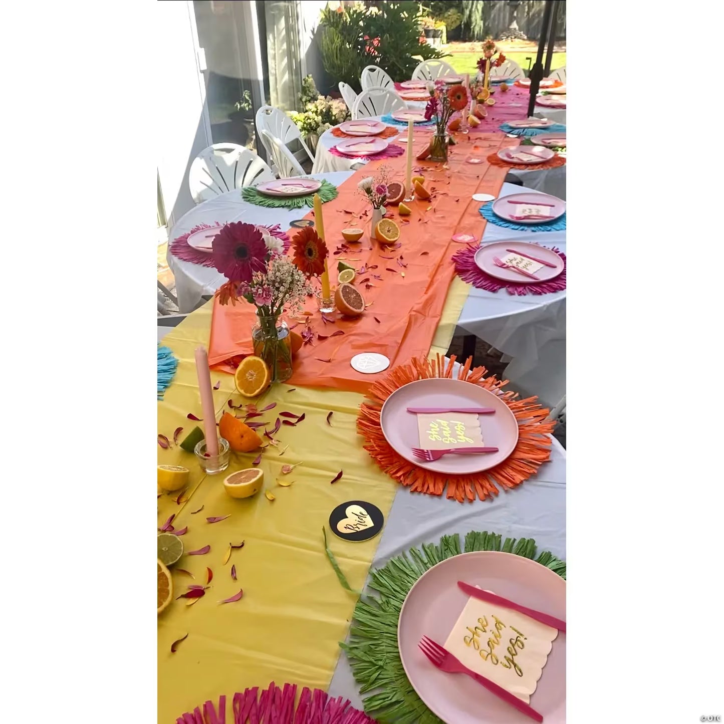 A long orange table set for a celebration with layered table runners in yellow, orange, and pink, decorated with candles, flowers, sliced oranges, and place settings with pink plates and gold calligraphy napkins.