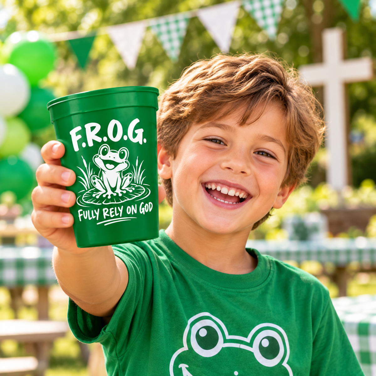 A smiling young boy in a green frog-print shirt holds up a green plastic cup printed with a frog graphic and the text F.R.O.G. fully rely on God themed cups.