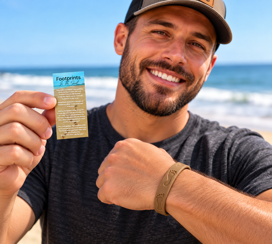 A smiling man on a beach holds up a small printed card while wearing a tan silicone wristband with footprints. The background shows the ocean and blue sky.