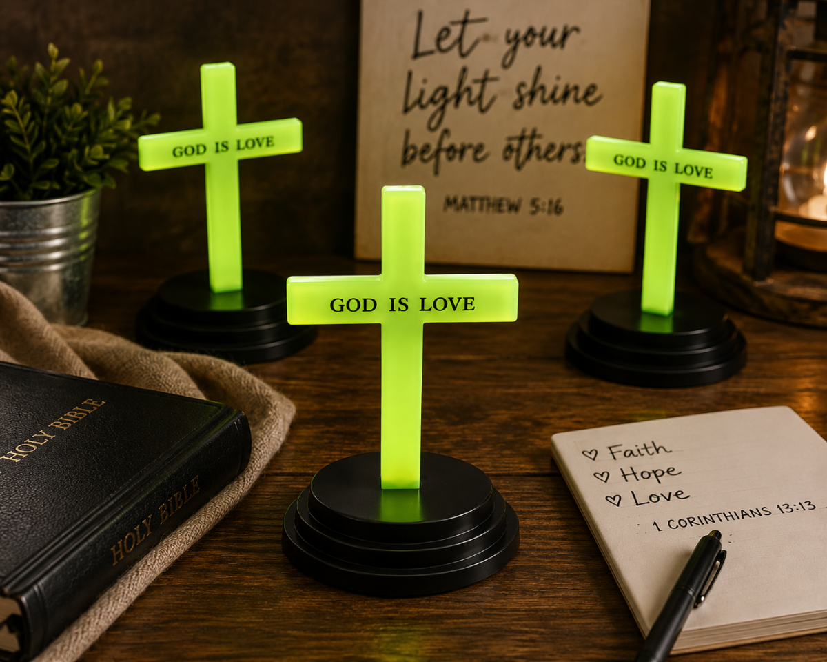 Three small glowing neon green cross candle holders with black bases, each engraved with 'GOD IS LOVE', displayed on a wooden table with a Bible, notebook, and decorative background elements.