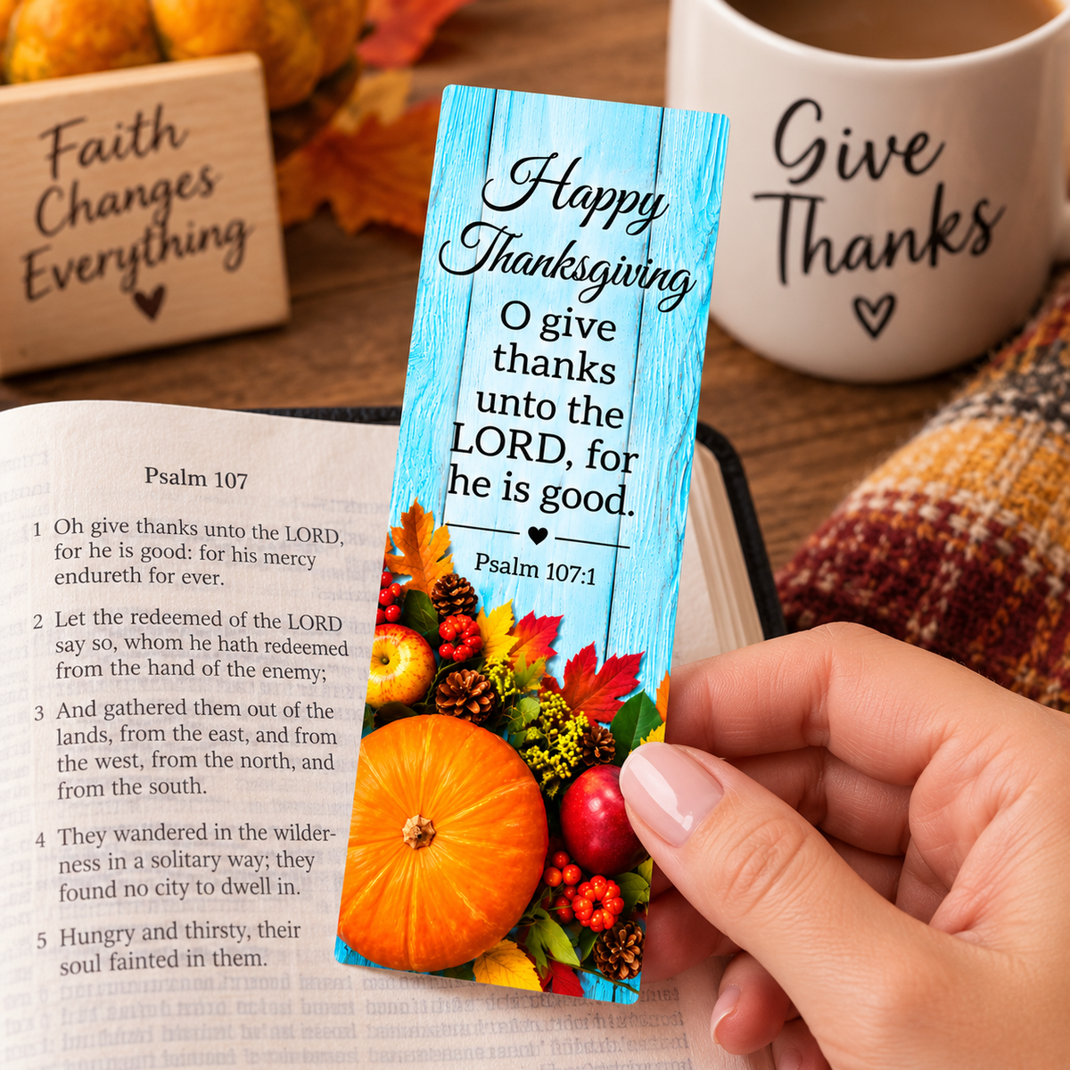 A hand holds a personalized Thanksgiving bookmark with a rustic blue wooden background, autumn leaves, pumpkins, and Psalm 107:1 text, set against an open Bible and a coffee mug.