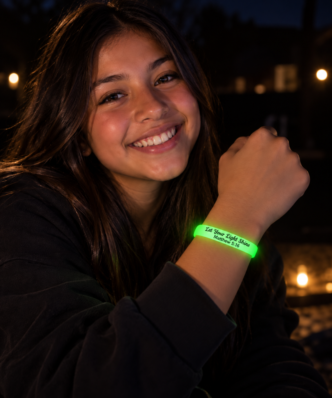 Teen girl smiling in the dark while wearing a glowing “Let Your Light Shine Matthew 5:16” bracelet on her wrist.