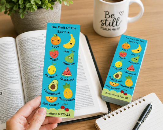 A hand holds a bright blue fruit of the spirit bookmark featuring cute cartoon fruits and words like Love, Joy, Patience, and Goodness, with Bible verse text. Another matching stack of bookmarks sits on a wooden desk beside an open book, a mug that says 'Be still,' and a notebook with a pen.