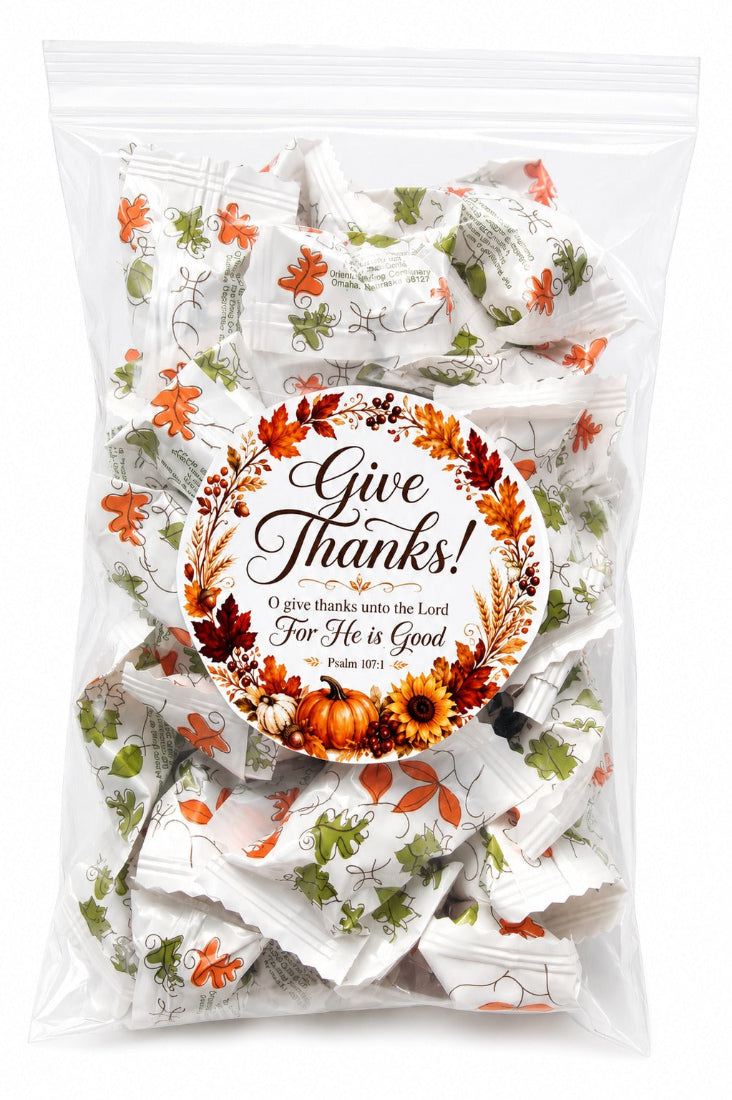 A clear plastic bag filled with individually wrapped white candies or chocolates, decorated with orange and green floral motifs, and featuring a round printed label that reads 'Give Thanks! O give thanks unto the Lord For He is Good Psalm 34:10' with autumn leaves and pumpkins.