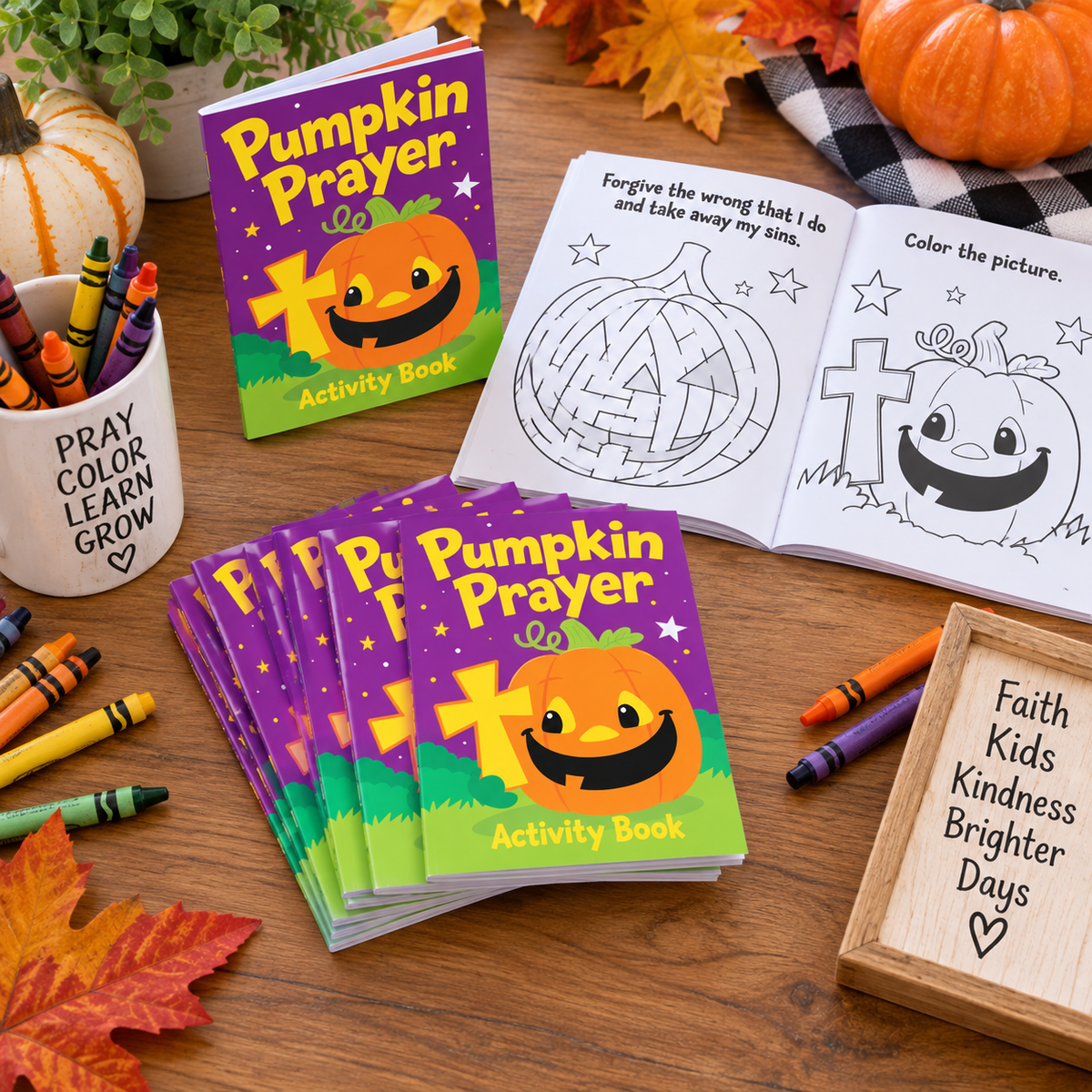 A set of 'Pumpkin Prayer' activity books displayed on a wooden table, alongside an open coloring book page, crayons, fall leaves, pumpkins, and a wooden sign.