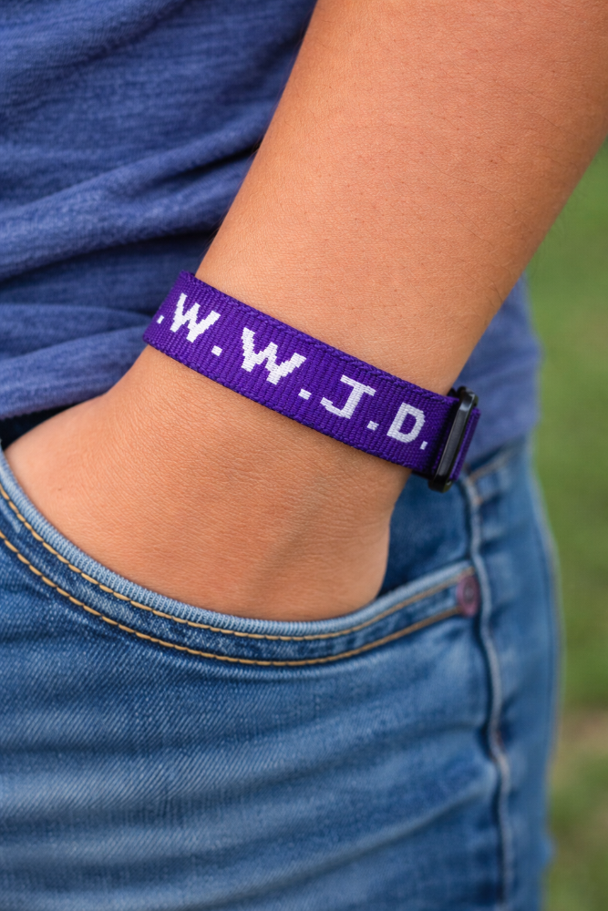Purple wristband with 'W.W.J.D.' on a person's wrist wearing blue jeans.