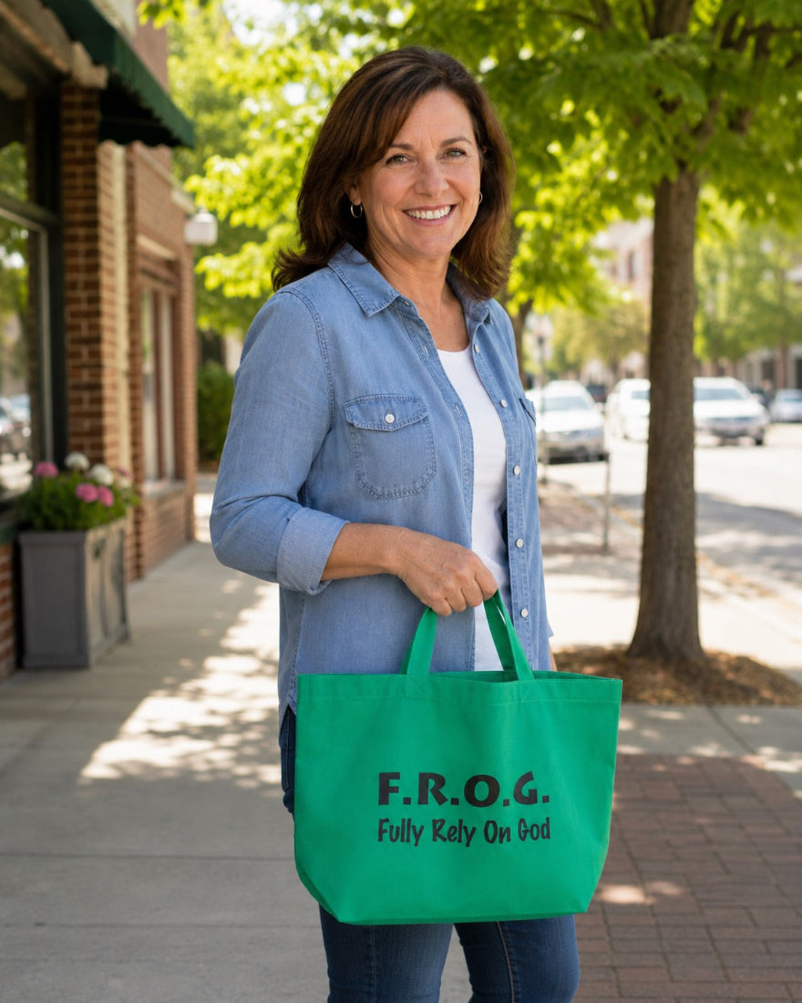 A smiling woman standing on a sidewalk outdoors holds a bright green tote bag printed with the text 'F.R.o.g. fully rely on God 