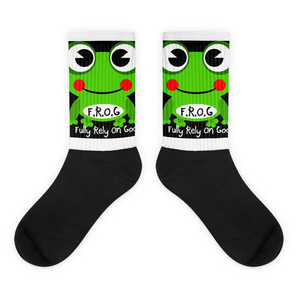 A pair of Christian crew socks on a white background featuring a smiling green frog with the text “F.R.O.G. – Fully Rely On God.” The socks have a white printed upper and a solid black foot, combining a fun design with a faith-based message.