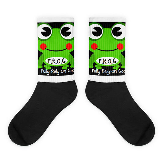 A pair of Christian crew socks on a white background featuring a smiling green frog with the text “F.R.O.G. – Fully Rely On God.” The socks have a white printed upper and a solid black foot, combining a fun design with a faith-based message.